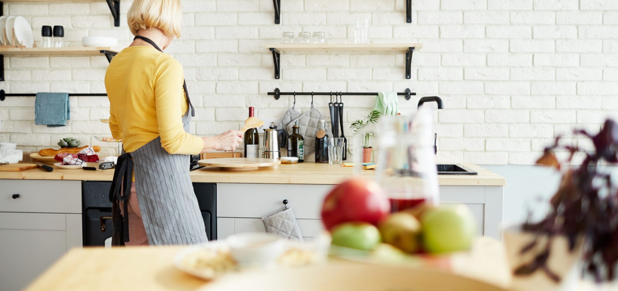Focus on blond-haired woman in stripped apron standing at kitchen counter and cooking food for dinner